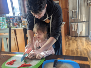A young kid learns to chop red bell pepper with the help of her parent for a delicious Shrimp and Grits recipe.
