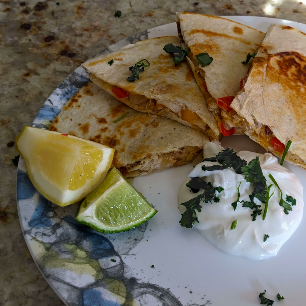A plate of golden-brown quesadillas with a side of sliced lemon and lime wedges. The quesadillas are stuffed with chunks of tilapia fillets, sliced red bell pepper, and onions. The dish is garnished with chopped cilantro and served on a white plate